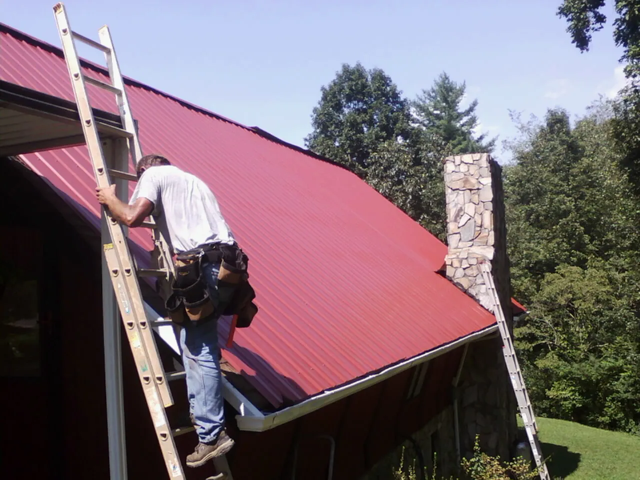 Roofer on a ladder beside a red metal roof and stone chimney — Soffit & Fascia Repair by Roofing Brothers in Mishawaka